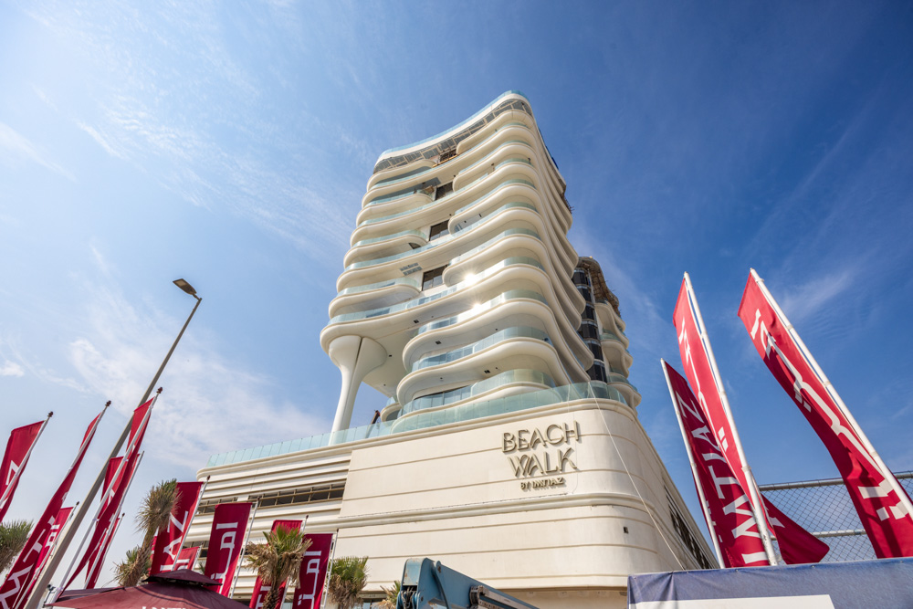 Beach Walk Residence Modern white building named Beach Walk under a blue sky, surrounded by palm trees and red flags.